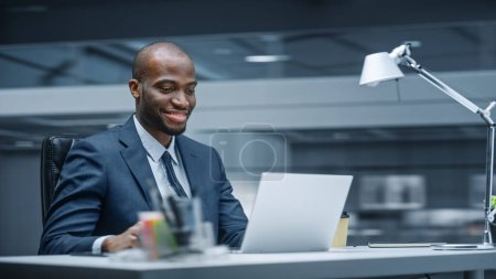 Téléchargez les photos : Bureau moderne : Homme d'affaires prospère assis au bureau en utilisant un ordinateur portable. Entrepreneur afro-américain en costume travaillant avec le marché boursier Investir - en image libre de droit
