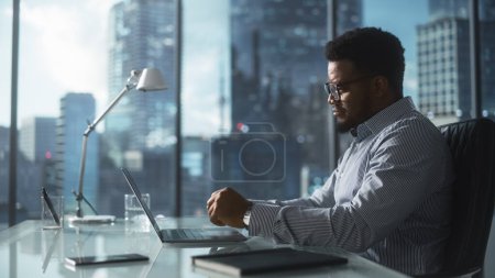 Téléchargez les photos : Homme d'affaires confiant en chemise rayée assis à un bureau dans un bureau moderne, en utilisant un ordinateur portable à côté de la fenêtre avec une grande ville avec gratte-ciel vue - en image libre de droit