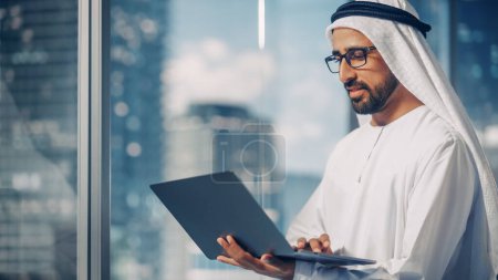 Foto de Empresario musulmán de éxito en la tradicional Kandura Blanca de pie en su oficina moderna, utilizando la computadora portátil junto a la ventana con rascacielos - Imagen libre de derechos