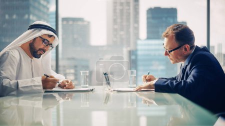 Foto de Dos hombres de negocios con diferentes nacionalidades sentados en la oficina y firmando contrato. Socio de negocios árabe hacer con el acuerdo financiero del inversor. Arabia Saudita - Imagen libre de derechos