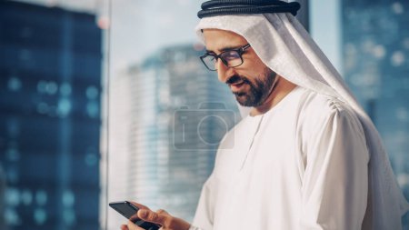Foto de Empresario musulmán exitoso en traje blanco tradicional parado en su oficina moderna, usando Smartphone al lado de la ventana con rascacielos. Exitoso. - Imagen libre de derechos