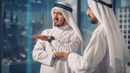 Téléchargez les photos : Deux hommes d'affaires musulmans prospères en tenue traditionnelle blanche debout au bureau Réunion, négociation et discussion sur les possibilités financières. Arabie saoudite - en image libre de droit