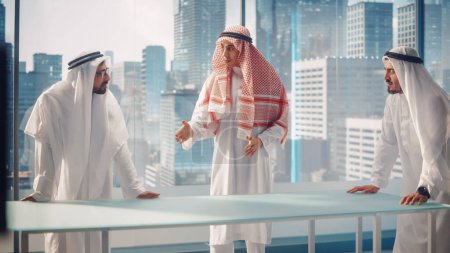 Photo for Three Successful Emirati Businessmen in White Traditional Kandura in Office Meeting, Negotiating and Talking About Financial Opportunities. Saudi - Royalty Free Image