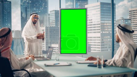 Foto de Emirati Businessman realiza una presentación de la reunión para socios comerciales. Arab Manager utiliza pizarra digital con pantalla verde vertical Mock Up Display - Imagen libre de derechos