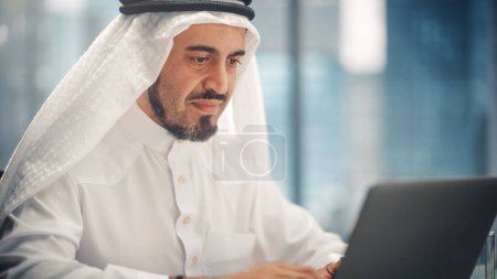 Téléchargez les photos : Homme d'affaires arabe prospère en tenue traditionnelle blanche assis au bureau et travaillant sur ordinateur portable. Gestionnaire des activités Planification Stratégie ministérielle - en image libre de droit
