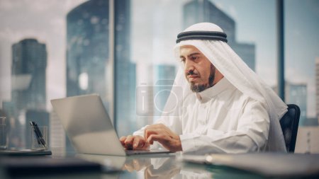 Téléchargez les photos : Homme d'affaires arabe prospère en blanc traditionnel Kandura assis au bureau et travaillant sur ordinateur portable. Gestionnaire des activités Planification des investissements - en image libre de droit