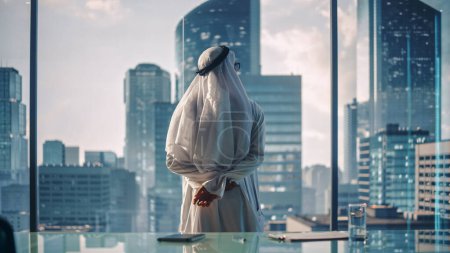 Téléchargez les photos : Homme d'affaires musulman prospère dans Kandura blanc traditionnel debout dans son bureau moderne regardant par la fenêtre sur la grande ville avec des gratte-ciel - en image libre de droit