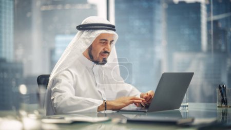 Téléchargez les photos : Homme d'affaires arabe en tenue traditionnelle blanche assis au bureau et travaillant sur ordinateur portable. Directeur des affaires Faire affaire avec succès d'investissement. Arabie saoudite - en image libre de droit
