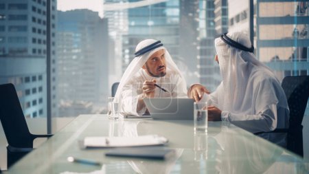 Foto de Dos exitosos hombres de negocios emiratíes en blanco Kandura tradicional sentados en la reunión de la oficina, negociando y hablando sobre oportunidades financieras - Imagen libre de derechos