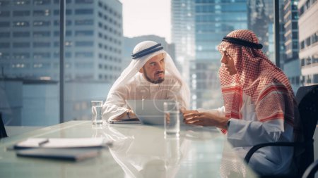Foto de Dos exitosos hombres de negocios emiratíes en blanco Kandura tradicional Sentado en la reunión de la oficina, negociando y hablando sobre oportunidades de inversión - Imagen libre de derechos