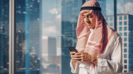 Téléchargez les photos : Jeune homme d'affaires musulman dans Kandura blanc traditionnel debout dans son bureau moderne, en utilisant Smartphone à côté de la fenêtre avec gratte-ciel. Succès - en image libre de droit