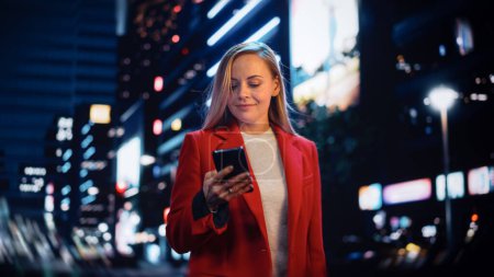 Téléchargez les photos : Portrait d'une belle femme en manteau rouge marchant dans une rue moderne avec des néons la nuit. Attrayant femelle en utilisant Smartphone et à la recherche - en image libre de droit