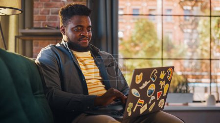 Foto de Hombre afroamericano guapo trabajando desde casa en la computadora portátil en Sunny Stylish Loft Office. Comprobación creativa masculina Redes sociales y navegación - Imagen libre de derechos