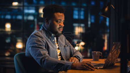 Téléchargez les photos : Jeune bel homme noir travaillant à domicile sur ordinateur portable dans un élégant appartement Loft le soir. mâle créatif obtient une excellente idée tout en vérifiant - en image libre de droit