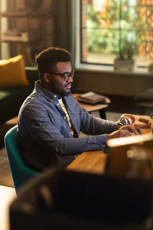 Vertikaler Bildschirm: Junger schöner schwarzer Mann, der tagsüber von zu Hause aus am Desktop-Computer in der stilvollen Loft-Wohnung arbeitet. Kreativer Männercheck