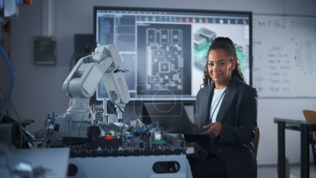 Téléchargez les photos : Portrait of Smiling Black Developer Using Laptop while Working with Robotic Arm in Modern University Classroom. Femme regardant la caméra. Succès et - en image libre de droit