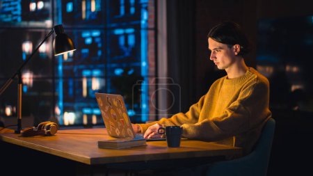Foto de Joven hombre guapo que trabaja desde casa en el ordenador portátil en el elegante apartamento Loft en la noche. Persona creativa que usa suéter amarillo acogedor y - Imagen libre de derechos
