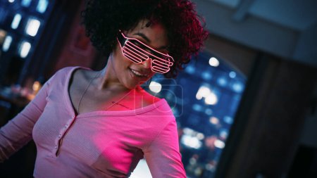 Foto de Primer plano Retrato de una joven mujer negra multiétnica bailando en gafas brillantes de neón futuristas, teniendo una fiesta en casa en Loft Apartment. Grabación - Imagen libre de derechos