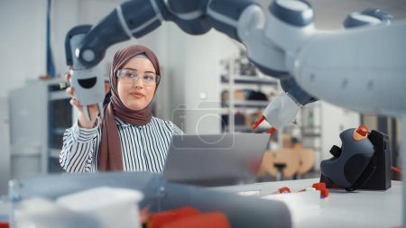 Foto de Modern Office: Portrait of Muslim Businesswoman Wearing Hijab and Working on Engineering Project, Coding on Laptop and Changing Robot Hand Position - Imagen libre de derechos
