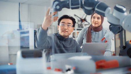 Foto de Arabic Female Future Engineer Looks At Work of Skilful Developer. Asian Man Explains Core Principles and Examins Robotic Arm. Analysis and Evaluation - Imagen libre de derechos