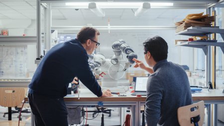 Foto de Adult Engineer Changes Position of Robotic Arm with Controller and Having a Discussion with Young Asian Developer. Startup Laboratory and Innovative - Imagen libre de derechos