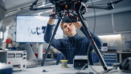 Téléchargez les photos : Jeune ingénieur masculin caucasien installe un processeur puissant dans le drone et les types sur ordinateur portable. Conception de véhicules aériens sans pilote dans un laboratoire moderne - en image libre de droit