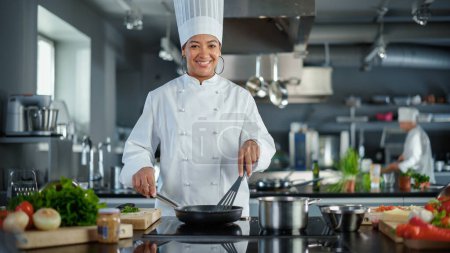 Foto de Restaurant Kitchen: Portrait of Black Female Chef in Action, Uses Pan to Cook Delicious, Traditional Authentic Food, Looks at Camera and Smiles - Imagen libre de derechos