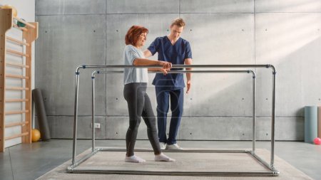 Téléchargez les photos : Physical Therapy Sports Center: Portrait of Strong Middle Aged Female Patient with Injury Successfully Walks Holding Parallel Bars. Physiotherapist - en image libre de droit