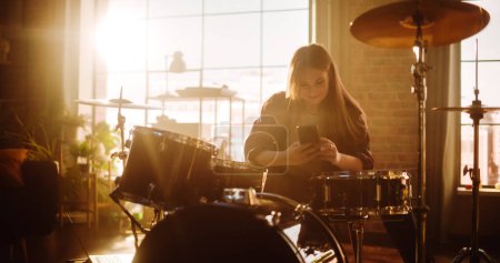 Téléchargez les photos : Young Female Pulls out Her Smartphone to Answer a Social Media Message. Expressive Drummer Finished Playing Drums in a Loft Music Rehearsal Studio - en image libre de droit