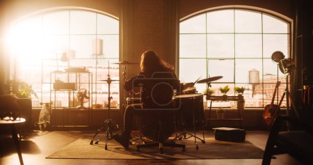 Téléchargez les photos : Person with Long Hair Sitting with Their Back to Camera, Playing Drums During a Band Rehearsal in a Loft Living Room with Warm Sunlight. Drummer - en image libre de droit