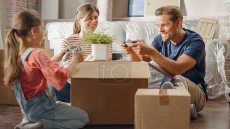 Téléchargez les photos : Happy Homeowners Moving In: Lovely Couple Sitting on the Floor of Cozy Apartment Unpacking Cardboard Boxes, Little Daughter Joins them. Cheerful Day - en image libre de droit