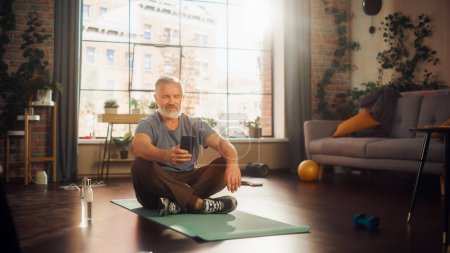 Foto de Strong Athletic Fit Middle Aged Man Using Smartphone in Between Workouts During Morning Exercises at Home in Sunny Apartment. Healthy Lifestyle - Imagen libre de derechos