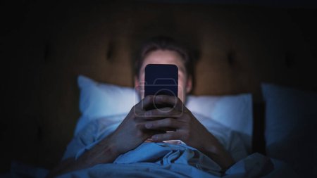 Hombre anónimo utiliza Smartphone en la cama en casa por la noche. Guapo Chico Navegando por las Redes Sociales, Leyendo Noticias, Haciendo Compras en Línea Tarde en la Noche. Centrarse en