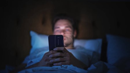 Homme caucasien utilise Smartphone au lit à la maison la nuit. Beau gars naviguant sur les médias sociaux, Lecture Nouvelles, Faire du shopping en ligne, Chatter avec des amis
