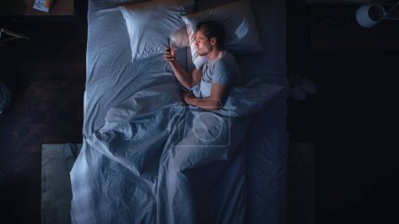 Schlafzimmer der Wohnung von oben: Schöner junger Mann, der nachts sein Smartphone im Bett benutzt. Guy surft durch soziale Medien, nutzt Dating-Apps, Remote-Arbeit