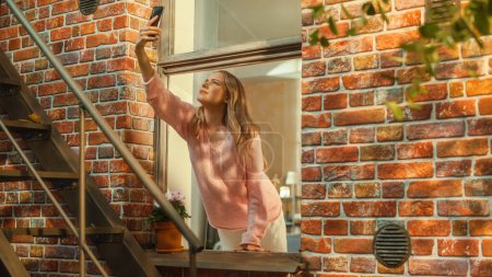 Genervte junge Frau auf der Suche nach einer mobilen Netzwerkverbindung auf ihrem Smartphone und greift mit dem Handy aus dem Fenster einer Wohnung in der Stadt