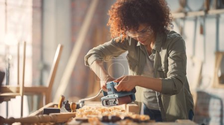 Female Carpenter Wearing Protective Safety Glasses and Using Electric Belt Sander to Work on a Wood Bar. Artist or Furniture Designer Working on a