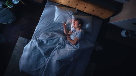 Top View Apartamento Dormitorio: Hombre joven guapo usando Smartphone en la cama por la noche. Chico Sonriente Navegando por Redes Sociales, Aplicaciones de Citas, Software de Trabajo Remoto
