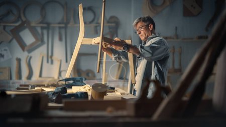 Carpintero adulto poniéndose gafas, leyendo planos y empezando a montar piezas de una silla de madera. Diseñador de Muebles Profesional Trabajando en un