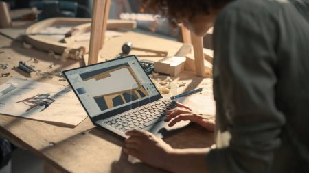 Téléchargez les photos : Plan rapproché d'un concepteur de meubles travaillant sur un ordinateur portable, créant un modèle numérique 3D d'une table de chevet en bois élégante. Personne créative - en image libre de droit