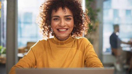 Téléchargez les photos : Portrait of an Attractive Arab Female Sitting at a Desk in Creative Agency. Young Stylish Manager with Curly Hair Smiling, Looking at Camera. Manager - en image libre de droit