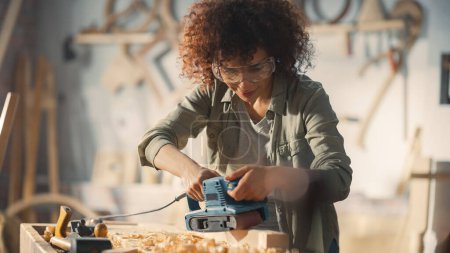 Multiethnic Woman Carpenter Wearing Protective Safety Glasses and Using Electric Belt Sander to Grind a Wood Block. Artist or Furniture Designer
