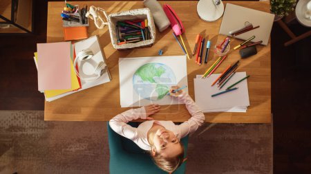 Téléchargez les photos : Vue du dessus : Petite fille dessinant notre belle planète Terre, levant les yeux et souriant. Enfant talentueux Imaginer notre planète natale comme un endroit heureux avec propre - en image libre de droit