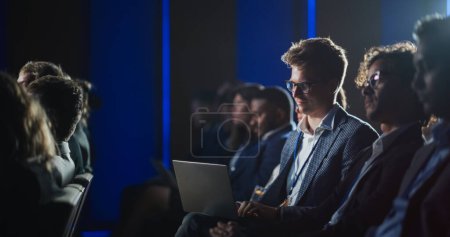 Téléchargez les photos : Young Man in Glasses Sitting in Crowded Audience at a Business Conference. Company Delegate Watching Inspirational Entrepreneurship Presentation About - en image libre de droit