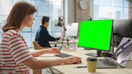 Téléchargez les photos : Portrait d'une jeune femme créative blanche travaillant sur un ordinateur à écran vert dans un bureau occupé. Femme Responsable Marketing Sourit tout en répondant à la clientèle - en image libre de droit
