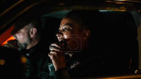 Porträt zweier Streifenpolizisten, die im Auto sitzen, essen und nachts über die Straßen schauen. Weiblicher schwarzer Polizist beißt beim Warten auf einen Donut