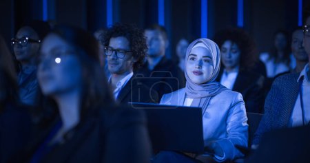 Photo for Arab Female Sitting in a Dark Crowded Auditorium at a Tech Conference. Young Muslim Woman Using Laptop Computer. Specialist in Hijab Watching - Royalty Free Image