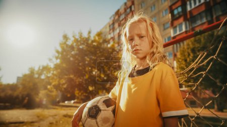 Porträt eines schönen blonden Mädchens in gelbem T-Shirt, das neben einem Tor in einem Hinterhof steht und einen Fußball hält. Junge Fußballspieler gesucht