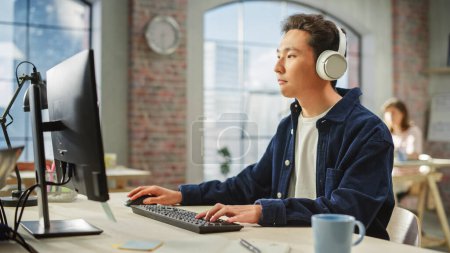 Portrait of Asian Creative Man Using Computer and Headphones in Bright Modern Office. Analyste de données masculin Développer une base de données tout en écoutant de la musique.