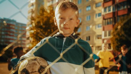 Téléchargez les photos : Portrait d'un beau garçon en sweat à capuche vert et blanc tenant un ballon de football dans le quartier. Jeune footballeur regardant la caméra, souriant. Les enfants - en image libre de droit